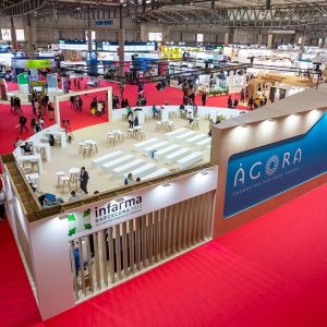 Stand de Infarma 2023 en una feria, con un diseño moderno y el logo de Infarma visible. El espacio está lleno de mesas y visitantes interactuando, mientras que se muestra un área de formación y un fondo vibrante con el logo del evento