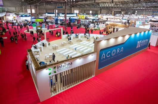 Stand de Infarma 2023 en una feria, con un diseño moderno y el logo de Infarma visible. El espacio está lleno de mesas y visitantes interactuando, mientras que se muestra un área de formación y un fondo vibrante con el logo del evento