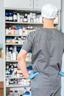 Farmacéutico de espaldas, vistiendo ropa de laboratorio y guantes azules, observando un estante lleno de frascos de medicamentos y productos farmacéuticos en una farmacia. La imagen refleja un entorno profesional en un espacio de trabajo farmacéutico.